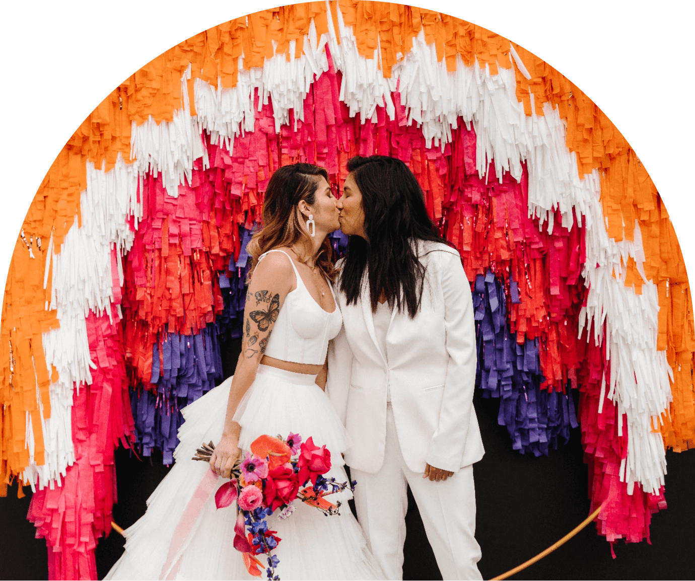 Two brides pose in front of a colorful streamer arch wedding photo backdrop.