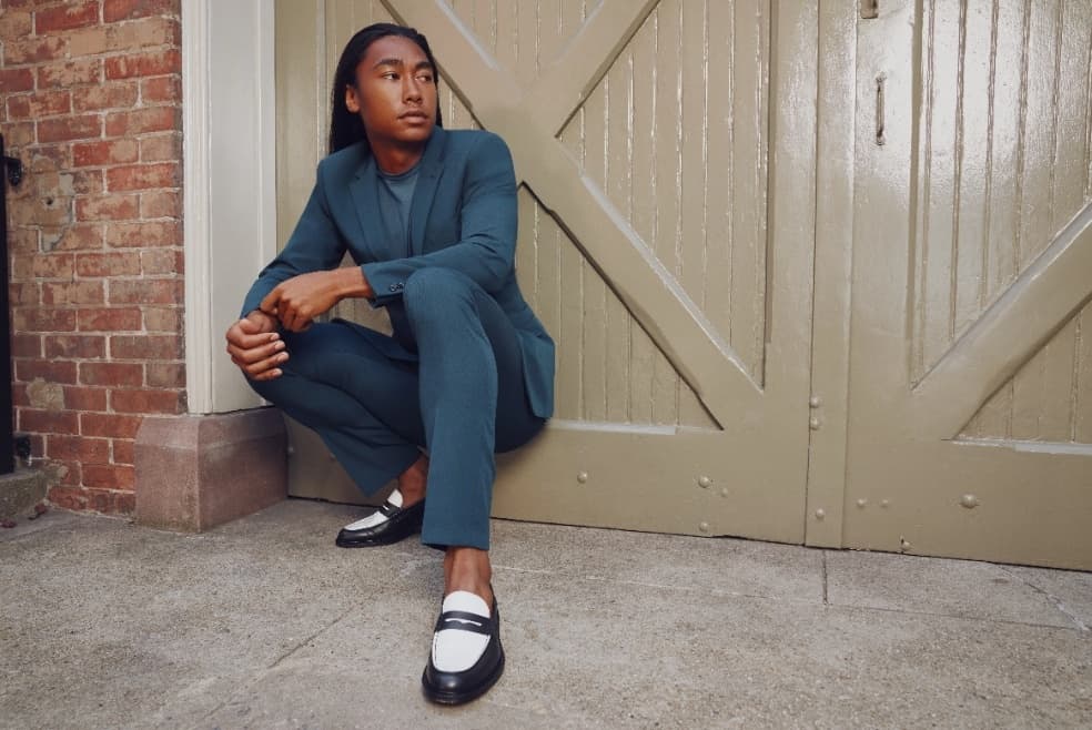 A man crouches coolly on the street in a teal suit with black and white loafers.