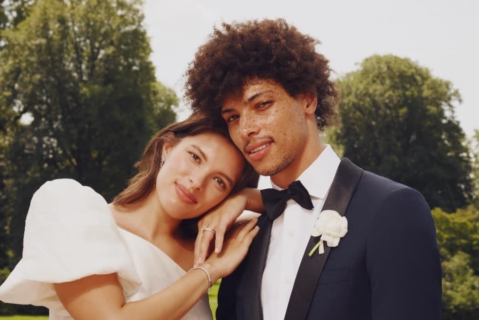 A bride and groom looking happily into the camera. A white boutonnière is pinned to the groom's lapel.