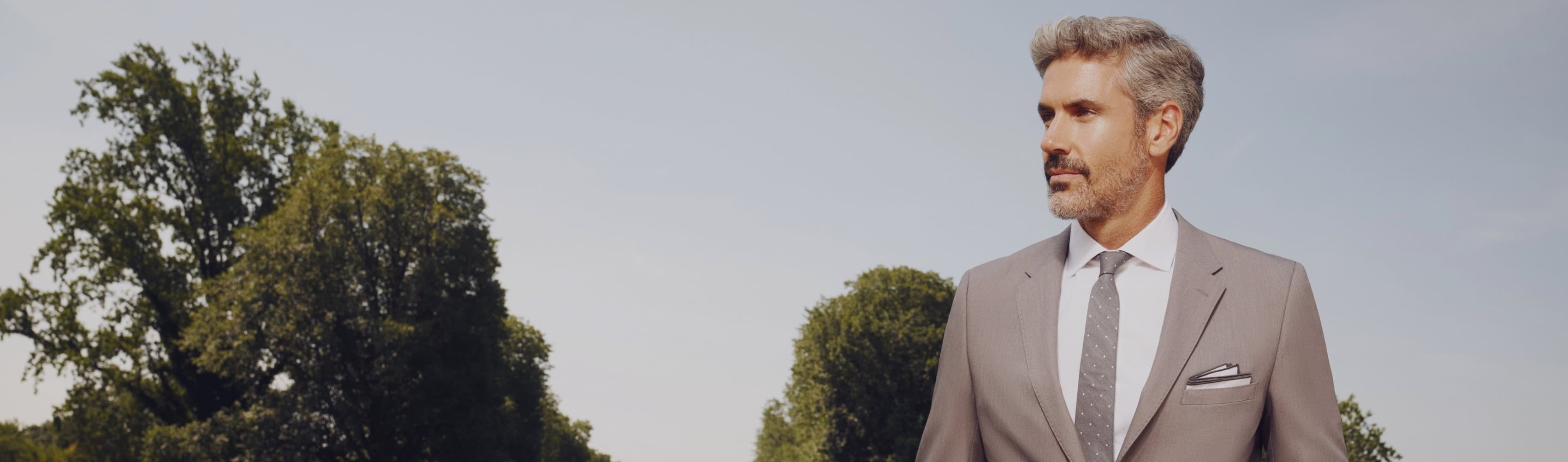 A handsome man in a stylish, well-fitted light gray suit gazes off camera.