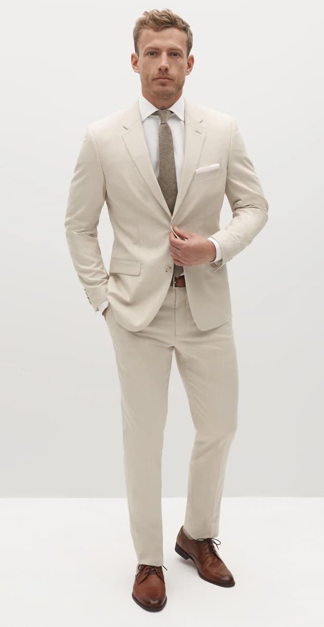 Slim man in light beige tan suit and brown accessories buttoning blazer jacket.